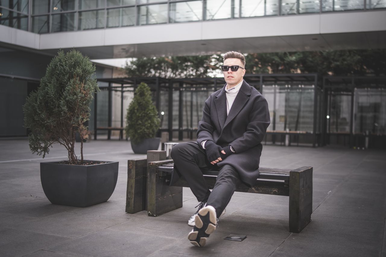 Stylish man with sunglasses and gloves sitting on a bench in an urban setting.