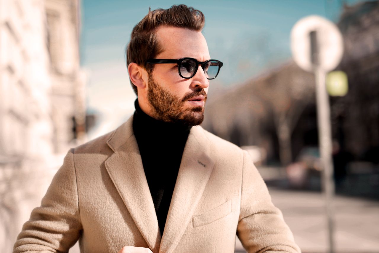 Stylish man in a beige coat and sunglasses looking away on a sunny city street.
